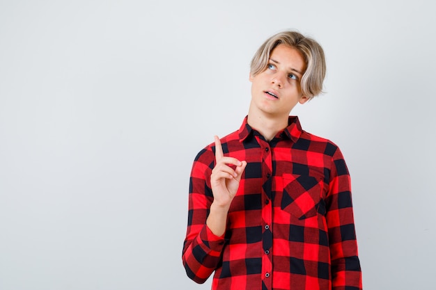 Foto junger teenager, der nach oben zeigt, im karierten hemd nach oben schaut und konzentriert aussieht, vorderansicht.