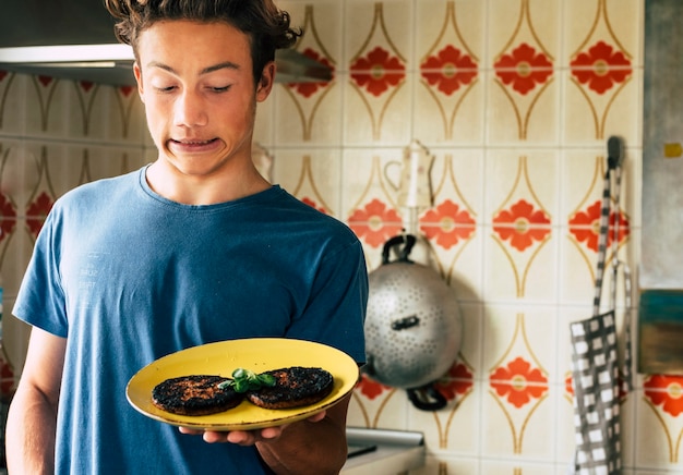 Junger Teenager mit verbranntem zu gekochtem Hamgurger mit lustigem Ausdruck in der Küche