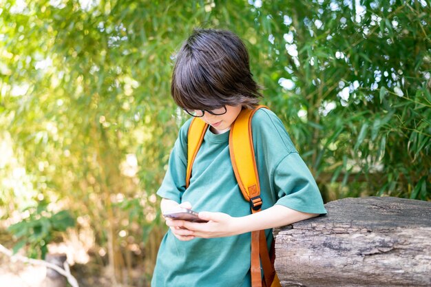 Junger Teensger-Junge mit Brille, der im Park bleibt und Nachricht auf seinem Smartphone-Kind mit Telefon im Freien im Sommer-Social-Media-Konzept tippt