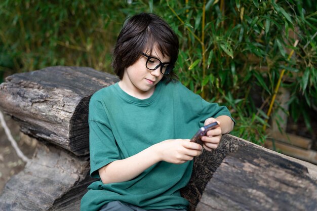 Junger Teensger-Junge mit Brille sitzt auf Öko-Bank im Park und tippt Nachricht auf seinem Smartphone-Kind mit Telefon im Freien im Sommer Social-Media-Konzept