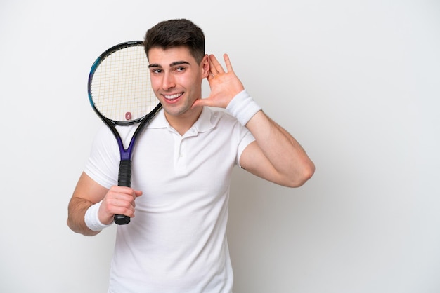 Junger Tennisspieler isoliert auf weißem Hintergrund, der etwas hört, indem er die Hand auf das Ohr legt