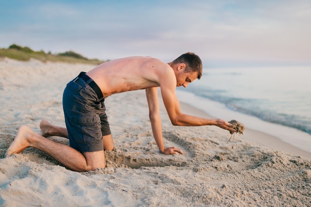 Junger topless barfüßiger Mann in kurzen Hosen, die auf Knien stehen und Grube durch Hände am Strand jenseits des Meeres im Sommerabend graben.
