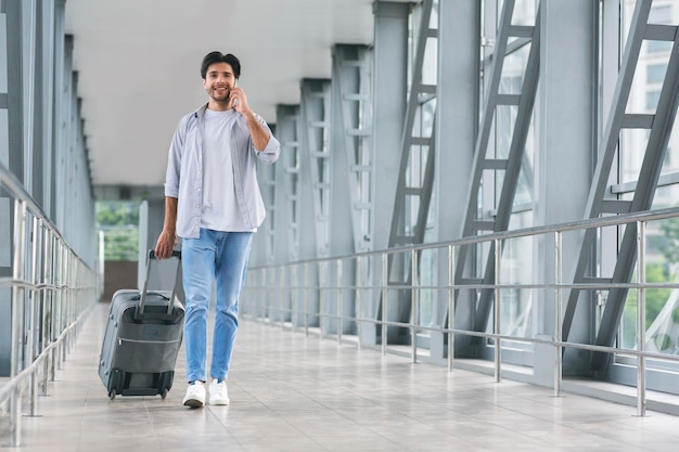 Junger Tourist, der am Telefon spricht und im Gepäck im Flughafen spazieren geht