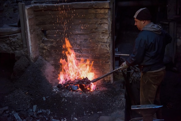 junger traditioneller Schmied, der mit offenem Feuer arbeitet Der Schmied macht Flammen in der Schmiede mit Funkenfeuerwerken, die heißes Eisen in der Werkstatt schmieden