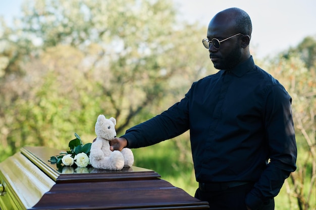 Junger trauernder afroamerikanischer Mann legt Teddybär auf den geschlossenen Sargdeckel