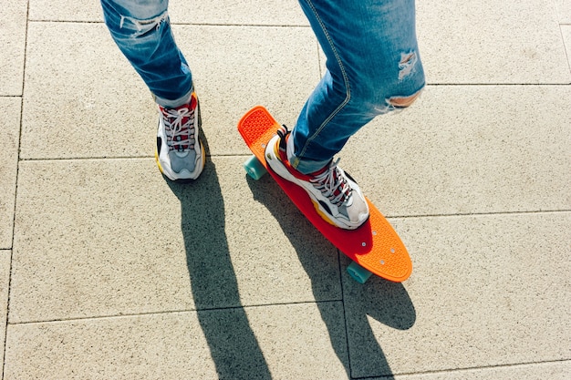 Junger Typ in zerrissenen Jeans und bunten Turnschuhen mit Penny Board auf dem Bürgersteig