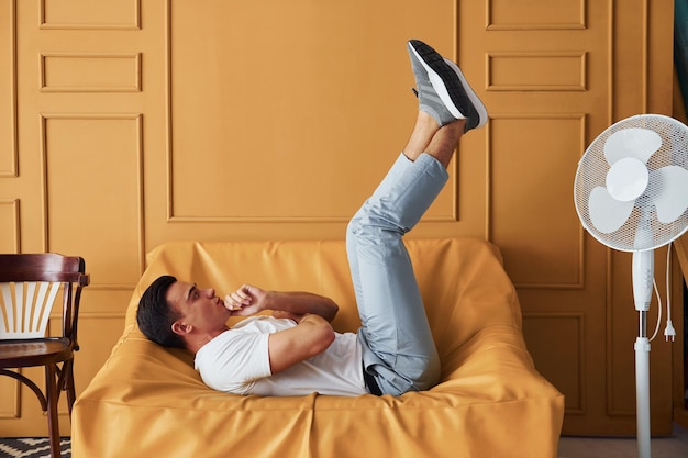 Junger Typ mit stylischem Haarschnitt und in Freizeitkleidung sitzt drinnen in einem Zimmer mit orangefarbenem Interieur