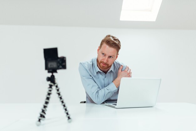 Junger überzeugter Mannsprecher, der auf dem Digitalkameraaufnahme vlog sitzt am Schreibtisch mit Laptop spricht.