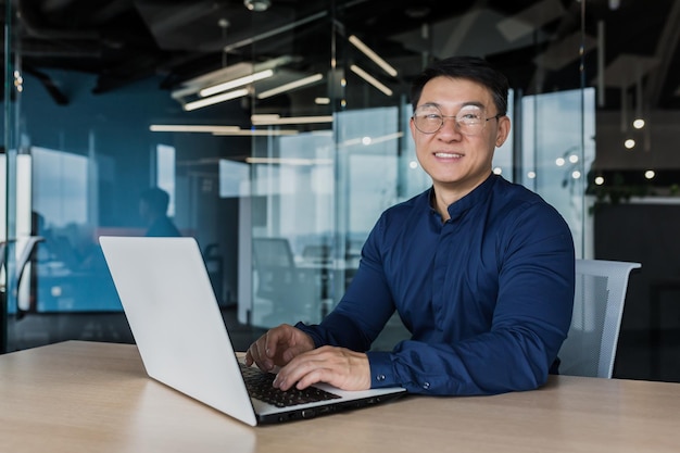 Junger und erfolgreicher asiatischer Geschäftsmann mit Brille, der an einem Laptop in einem modernen Bürogebäude arbeitet