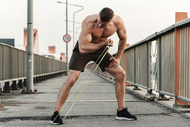 Junger und muskulöser Mann während des Trainings mit Widerstandsgummibändern auf einer Straße. Bizeps-Curls-Übung.