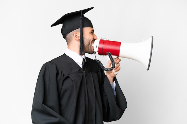 Junger Universitätsabsolvent vor isoliertem weißem Hintergrund, der durch ein Megaphon schreit