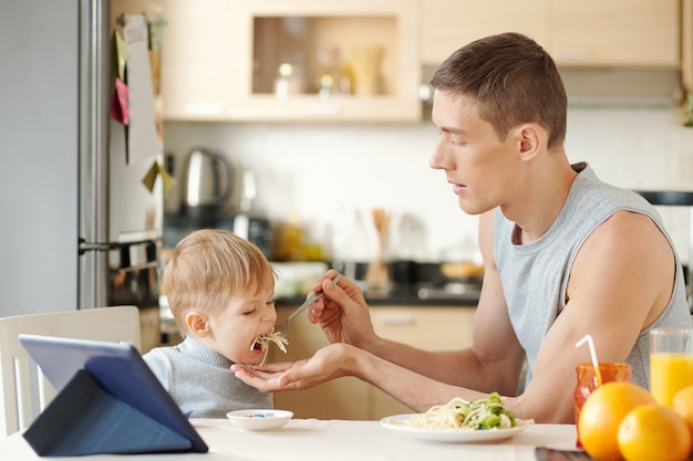 Junger Vater füttert seinen kleinen Sohn während des Mittagessens in der Küche