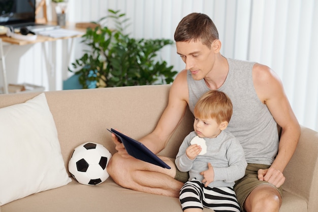 Junger Vater sitzt mit seinem kleinen Sohn auf dem Sofa und sieht sich gemeinsam Cartoons auf einem digitalen Tablet an