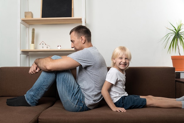 Junger Vater sitzt Rücken an Rücken mit kleinem Sohn auf dem Sofa und ignoriert sich gegenseitig Kind macht Vater wütend Generationskonflikt
