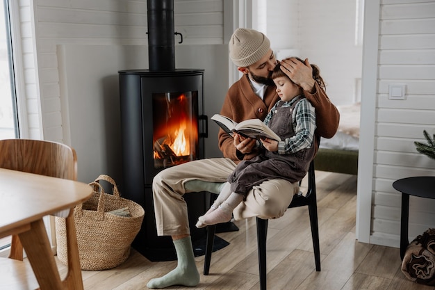 junger Vater und kleine Tochter in den Armen lesen ein Buch zusammen in der Nähe eines warmen Kamin in einem weißen Holzhaus