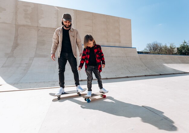 Foto junger vater und sein sohn fahren skateboards in einem skatepark mit rutschen draußen am sonnigen tag.