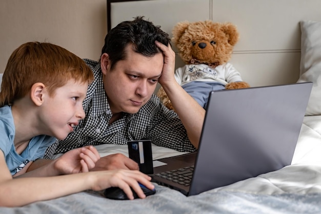 Junger Vater und Sohn, die zusammen mit der Familie online einkaufen. Kind, rothaariger Junge und Vater am Laptop, wählen Einkäufe, die der Mann dachte, Internet-Zahlung