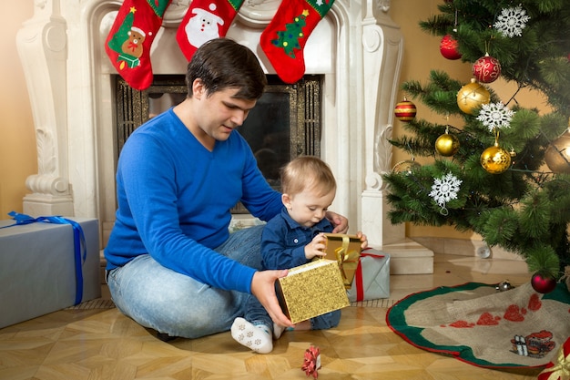 Junger Vater und süßer Babysohn öffnen Weihnachtsgeschenke auf dem Boden im Wohnzimmer