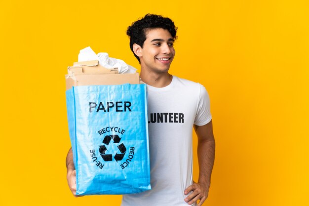 Junger venezolanischer Mann, der eine Recycling-Tasche voll Papier hält, um das Aufstellen mit Armen an der Hüfte und lächelnd zu recyceln