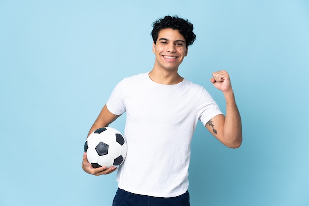 Junger venezolanischer Mann lokalisiert auf blauem Hintergrund mit Fußball, der einen Sieg feiert