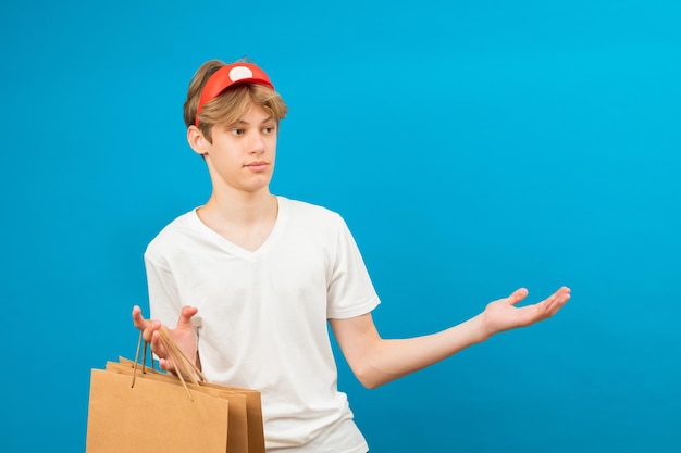 Junger Verkäufer in lässigem weißem T-Shirt mit Papiertüte isoliert auf blauem Hintergrund Studioporträt People Lifestyle-Konzept Mock-up-Kopierbereich Teen holding Paketbeutel mit Einkäufen