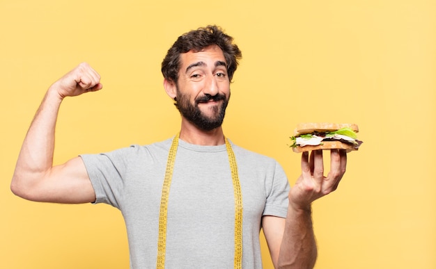 Junger verrückter bärtiger Mann, der glücklichen Ausdruck nährt und ein Sandwich hält