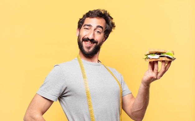 Junger verrückter bärtiger Mann, der glücklichen Ausdruck nährt und ein Sandwich hält