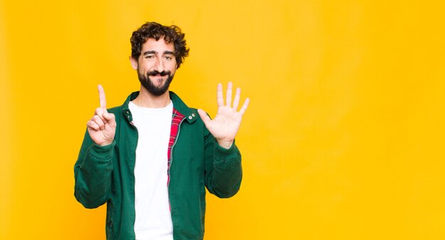 Junger verrückter bärtiger Mann, der lächelt und freundlich aussieht, Nummer sechs oder Sechste mit der Hand nach vorne zeigend, gegen flache Wand herunterzählend