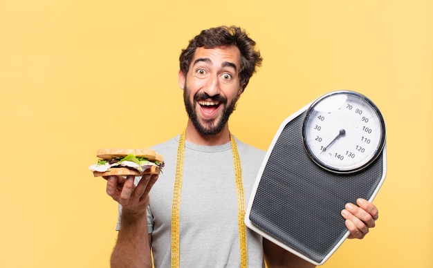 Junger verrückter bärtiger Mann, der überraschten Gesichtsausdruck nährt und eine Waage und ein Sandwich hält