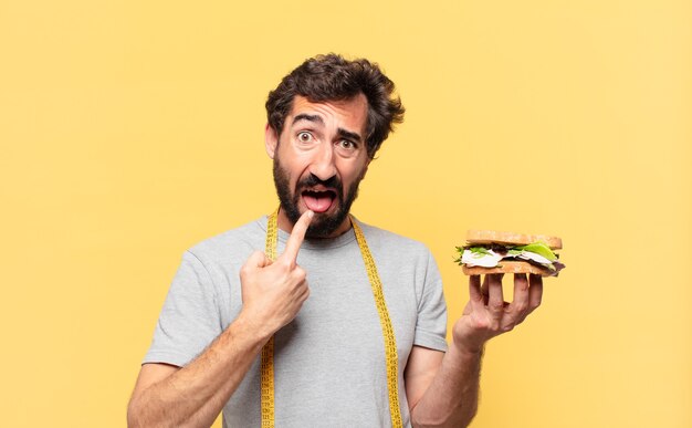 Junger verrückter bärtiger Mann, der wütenden Ausdruck nährt und ein Sandwich hält