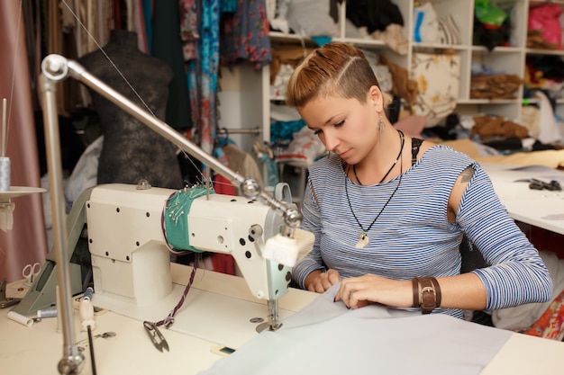 Junger weiblicher blonder Damenschneiderinstoffdesigner arbeitet an Nähmaschine. In ihrem Arbeitsbereich.