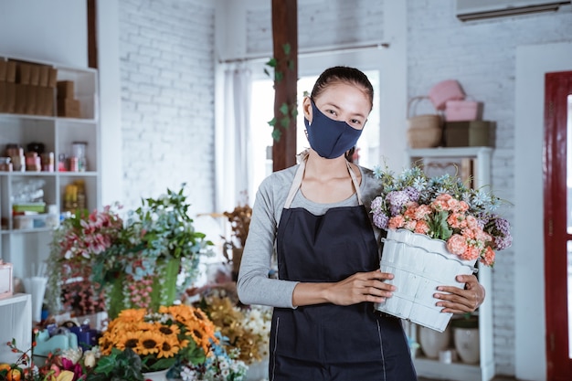 Junger weiblicher Florist, der Schürze und Gesichtsmaske hält Eimerblume lächelnd betrachtet Kamera