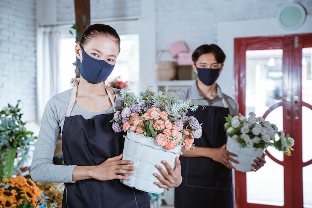 Junger weiblicher Florist, der Schürze und Gesichtsmaske hält Eimerblume lächelnd betrachtet Kamera