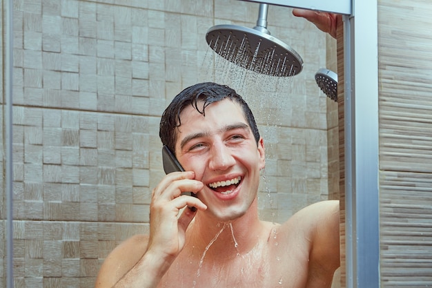 Junger weißer Mann lacht, während er am Telefon spricht und unter Wasserstrahlen steht, die von der Dusche laufen.