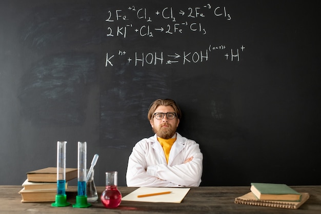 Junger zeitgenössischer Chemielehrer im Weißmantel, der seine Arme auf der Brust während des Online-Unterrichts an der Tafel mit chemischen Formeln kreuzt