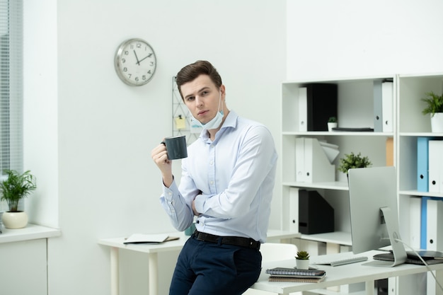 Junger zeitgenössischer Geschäftsmann mit einer Tasse Tee oder Kaffee, der während der Pause an seinem Arbeitsplatz mit Computer vor der Kamera steht