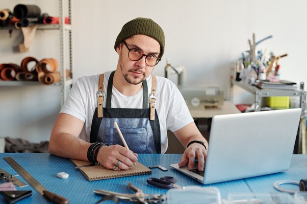 Junger zeitgenössischer Lederarbeiter in der Arbeitskleidung, die Skizze von neuen Gegenständen beim Sitzen am Tisch in der Werkstatt macht