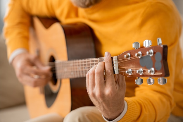 Junger zeitgenössischer männlicher Musiker in Freizeitkleidung, der Akustikgitarre spielt, während Online-Kurs des Musikmachens zu Hause nimmt