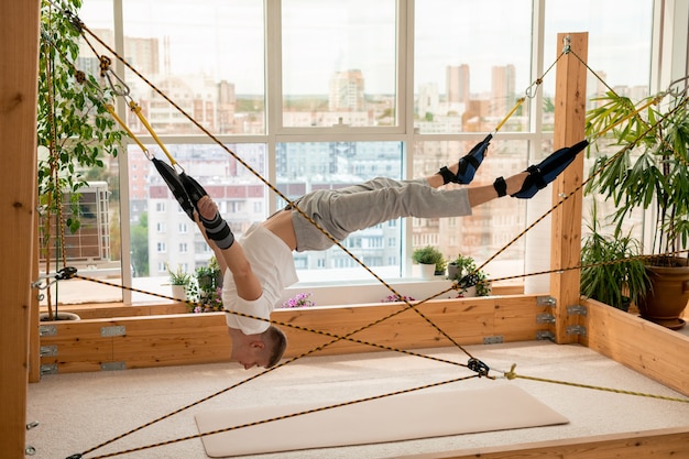 Junger zeitgenössischer Sportler, dessen Arme und Beine an Seilen befestigt sind, die schwierige Luft-Yoga-Übungen machen, während sie über dem Boden hängen