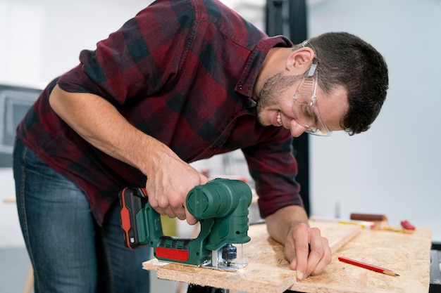 Junger Zimmermann schneidet eine OSB-Platte mit den Händen eines Puzzle-Handwerkers in Schutzhandschuhen aus Stoff