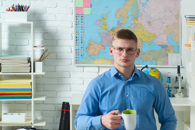 Junger zufälliger Mann mit Brille, der am Schreibtisch sitzt und mit einer Tasse Kaffee studiert.