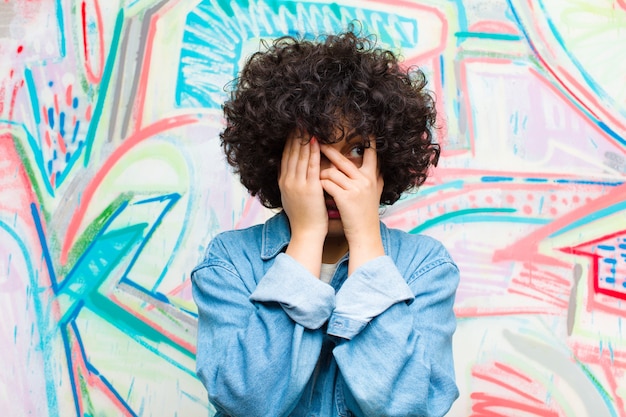 Junges Afrofrauen-Bedeckungsgesicht mit den Händen, spähend zwischen Finger mit überraschtem Ausdruck und schauen zur Seite auf Graffitiwand