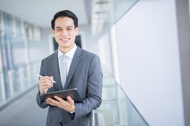 Junges asiatisches Geschäftsmannlächeln, das eine Tablette am intelligenten Geschäftskonzept des Bürobereichsmannes hält