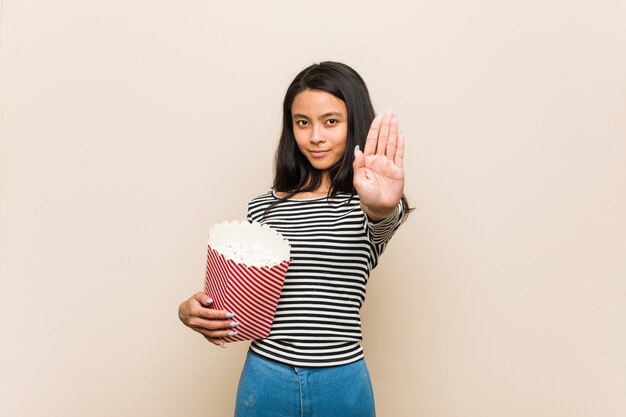 Junges asiatisches Mädchen, das einen Popcorneimer steht mit der ausgestreckten Hand zeigt das Stoppschild und verhindert Sie hält.