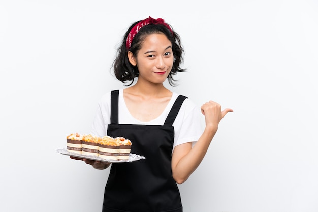 Junges asiatisches Mädchen, das viele Muffinkuchen über der lokalisierten weißen Wand zeigt auf die Seite hält, um ein Produkt darzustellen