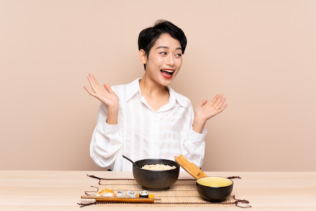 Junges asiatisches Mädchen in einer Tabelle mit Schüssel Nudeln und Sushi mit Überraschungsgesichtsausdruck