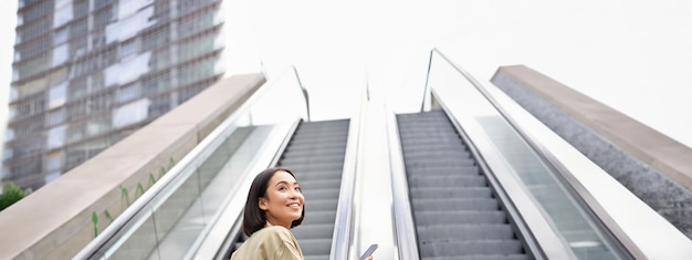 Junges asiatisches Mädchen steigt auf einer Rolltreppe hinauf und hält lächelnd ein Smartphone in der Hand, während es durch die Stadt geht