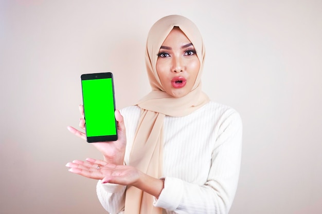 Junges asiatisches muslimisches schönes Mädchen mit Kopftuch ist schockiert und zeigt Greenscreen-Smartphone