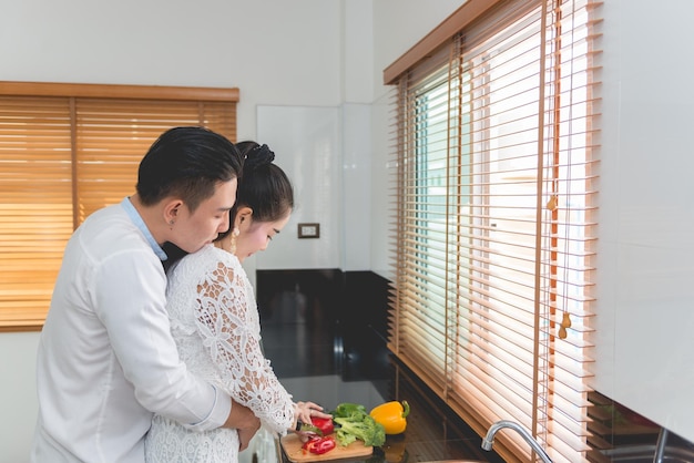 Junges asiatisches Paar, das zusammen mit Loveshe kocht, schneidet Gemüse, das er von hinten umarmt
