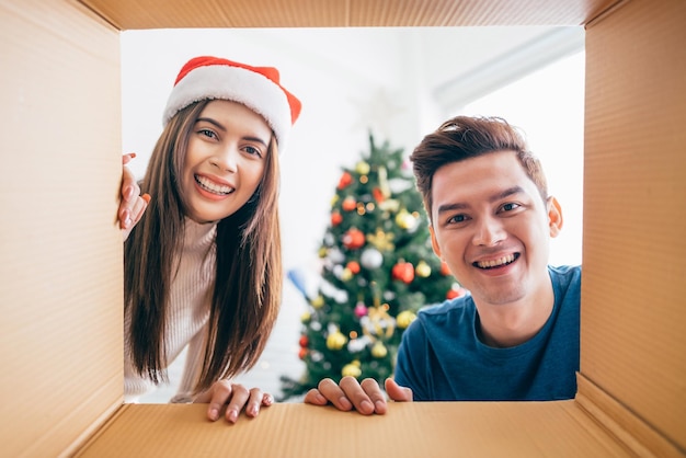 Junges asiatisches Paar öffnet ein Weihnachtsgeschenk zusammen mit einem Lächeln im Inneren der Schachtel mit einem geschmückten Weihnachtsbaum im Hintergrund Boxing Day während der Weihnachtsferien